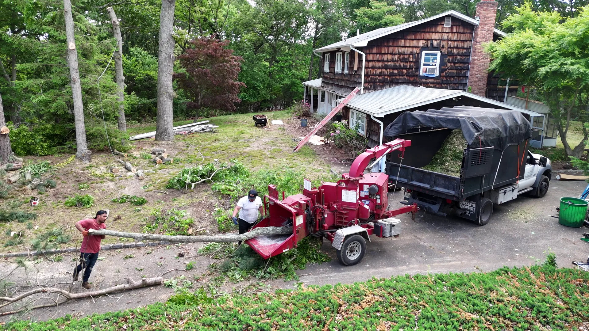 Aerial view of a bright red wood chipper and crew working a wooded driveway, chips feeding out.